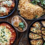 A dining table featuring chicken parmesan with spaghetti and eggplant parmesan with salad, styled with rustic cutlery and warm lighting.