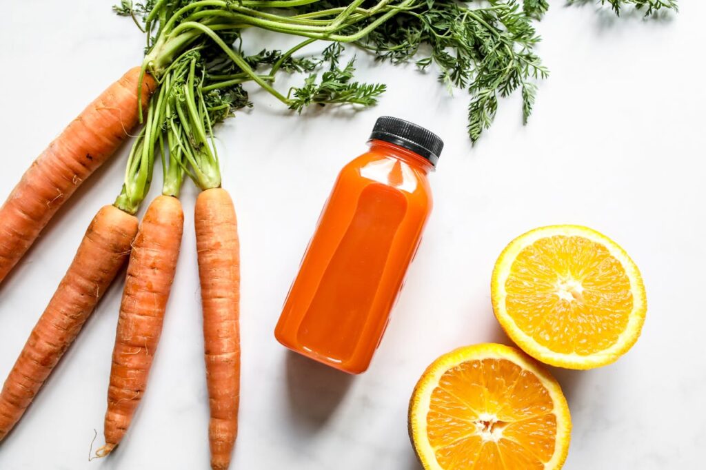 Fresh carrots, a bottle of carrot juice, and sliced oranges on a white background, highlighting the natural ingredients for carrot juice preparation.