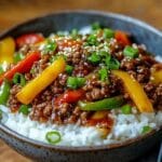 Authentic Japanese Pepper Steak Recipe for Beginners 7 A steaming bowl of beef and pepper rice, featuring juicy caramelized ground beef in a savory soy-based sauce, mixed with vibrant red, yellow, and green bell peppers. Served over fluffy white rice, garnished with sesame seeds and chopped green onions, with a pair of black chopsticks resting on the bowl. Warm, inviting lighting enhances the dish's rich colors and textures.