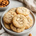 Almond Cookies Recipe: The Ultimate Nutty Delight 4 A close-up of almond cookies recipe featuring golden cookies topped with sliced almonds, stacked on a ceramic plate with whole almonds in the background.