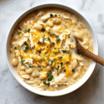 Creamy White Chicken Chili Recipe – Easy, Hearty, and Comforting 7 A close-up of creamy white chicken chili in a bowl with a wooden spoon lifting a scoop. Loaded with shredded chicken, white beans, cheddar cheese, corn, and garnished with parsley.