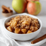 Sourdough Discard Apple Crisp: A Tangy Twist on a Classic Fall Dessert 7 Close-up of sourdough discard apple crisp in a white bowl with oat crumble topping, cinnamon apples, and blurred cinnamon sticks in the background.