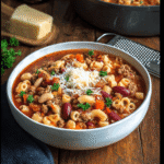 A bowl of Copycat Olive Garden Pasta e Fagioli topped with Parmesan cheese and parsley, surrounded by a cheese wedge and grater on a rustic table.