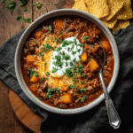 Bowl of hearty pumpkin chili topped with sour cream, cheddar, and parsley, served with tortilla chips on a wooden table.