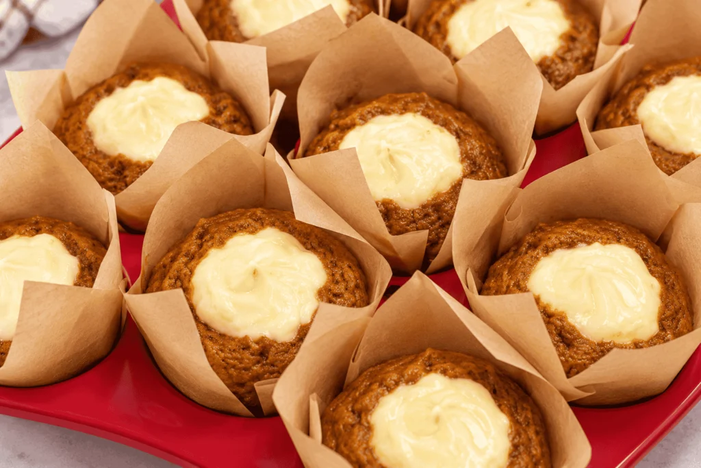 Freshly baked pumpkin cream cheese muffins with golden-brown pumpkin spice tops and rich, creamy cheesecake centers, nestled in brown parchment muffin liners.