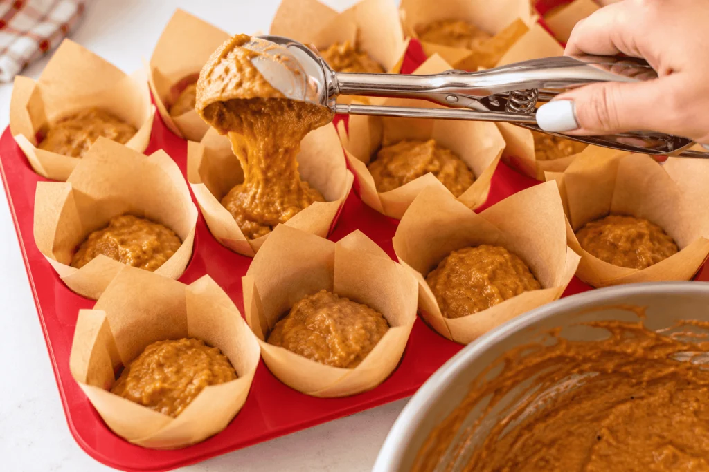 Homemade pumpkin cream cheese muffins being prepared with a cookie scoop, filling parchment-lined muffin cups with spiced pumpkin batter in a red muffin tray.