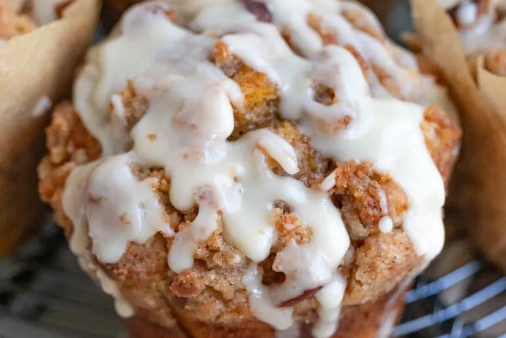 Close-up of a golden pumpkin streusel muffin with pecan topping, covered in thick vanilla glaze and resting on a cooling rack.